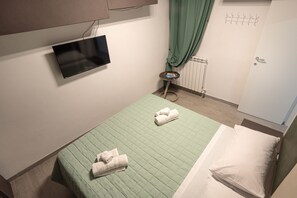 Quarto pequeno com uma cama de solteiro, colcha verde, televisão de tela plana na parede, uma mesa com um livro e um radiador.