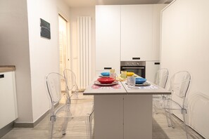 Een kleine keuken met witte kastjes, een centraal keukenblok met een tafel, en transparante stoelen.