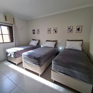 A room with three beds aligned against the wall, a window with curtains, and a wall-mounted air conditioning unit. - CASA 1111 FUNDO (São José do Rio Preto)