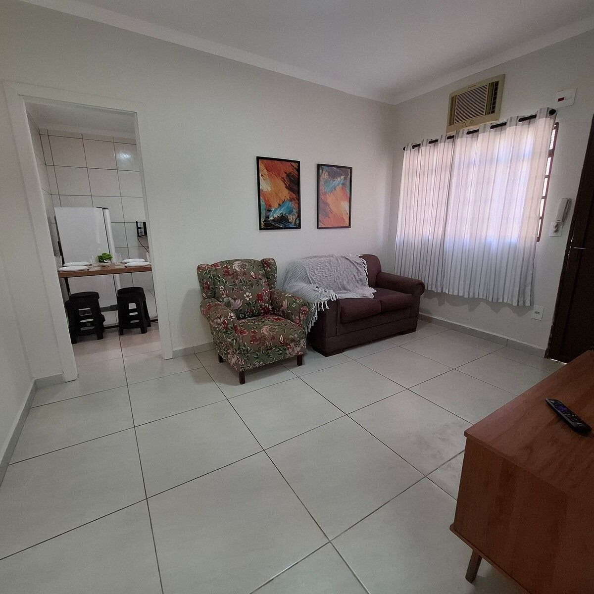 A living room with a sofa, a floral armchair, a dining table, and a kitchen area visible through an open doorway.