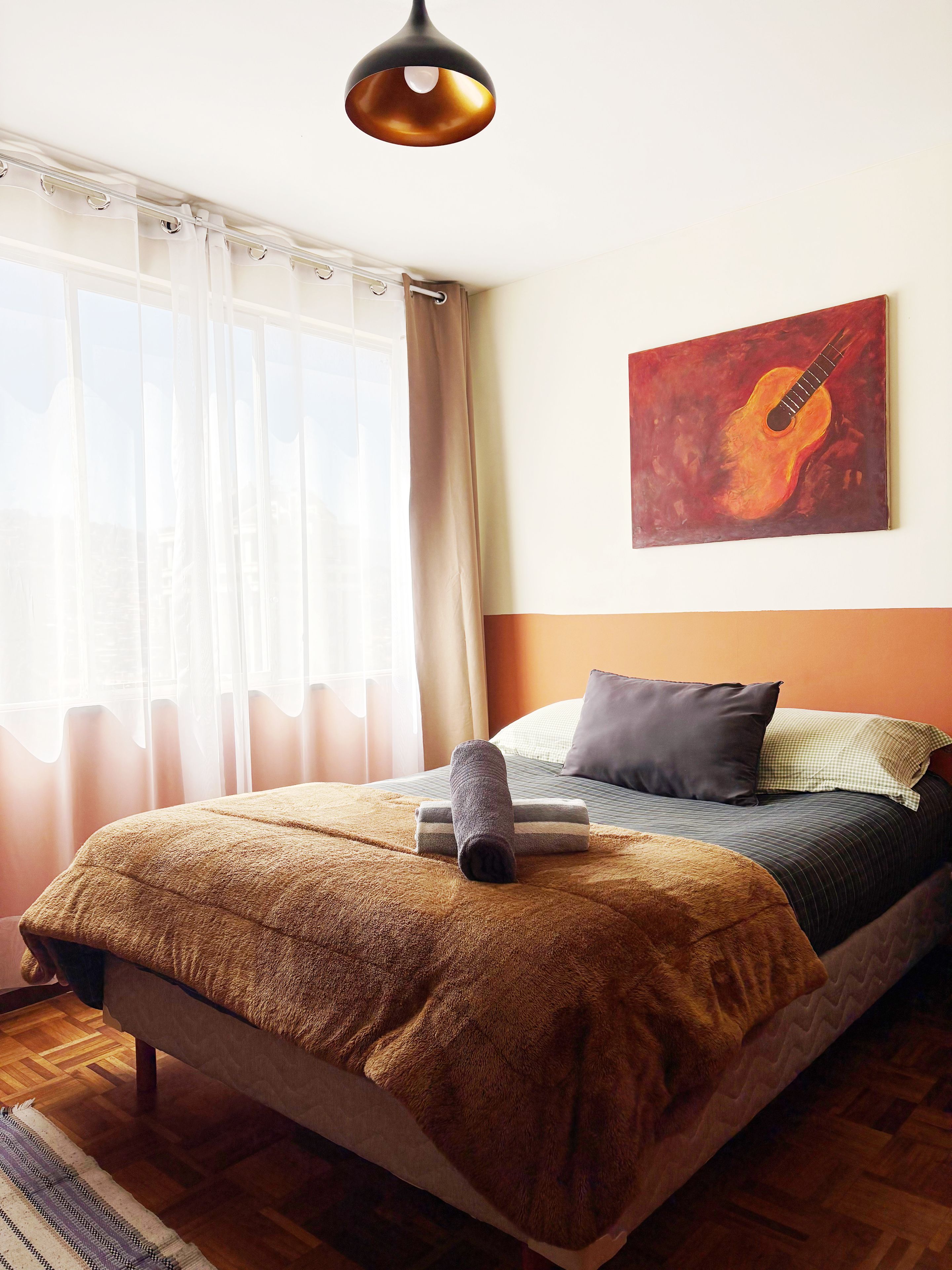 Une chambre à coucher avec un lit, un tableau représentant une guitare et une fenêtre avec des rideaux.