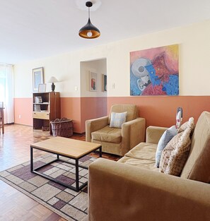 Uma sala de estar com sofá, poltronas, mesa de centro, luminária, pintura e estante de livros.