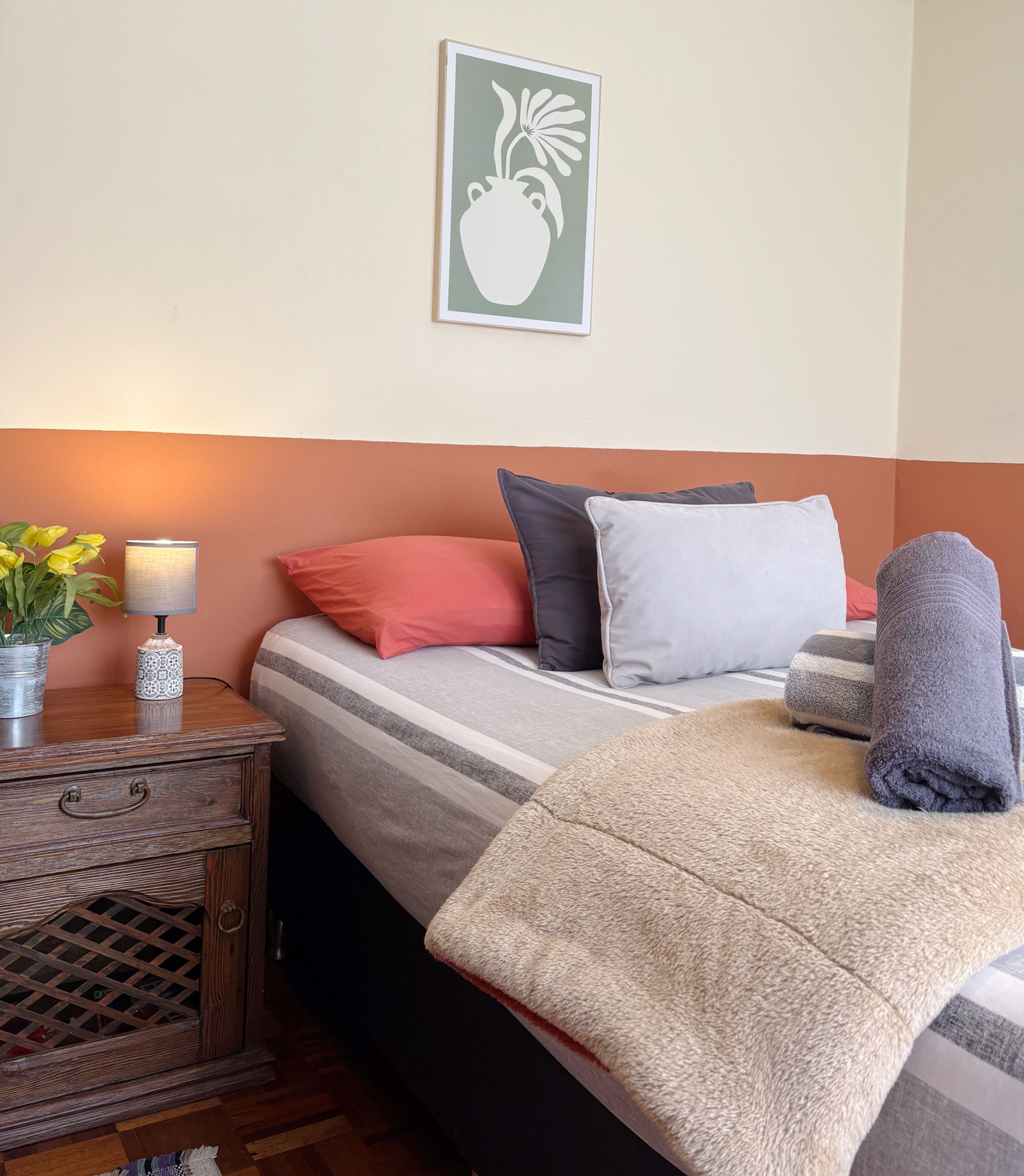 Habitación de hotel ordenada con cama, almohadas, lámpara de noche, una mesita con un jarrón de flores y un cuadro en la pared.