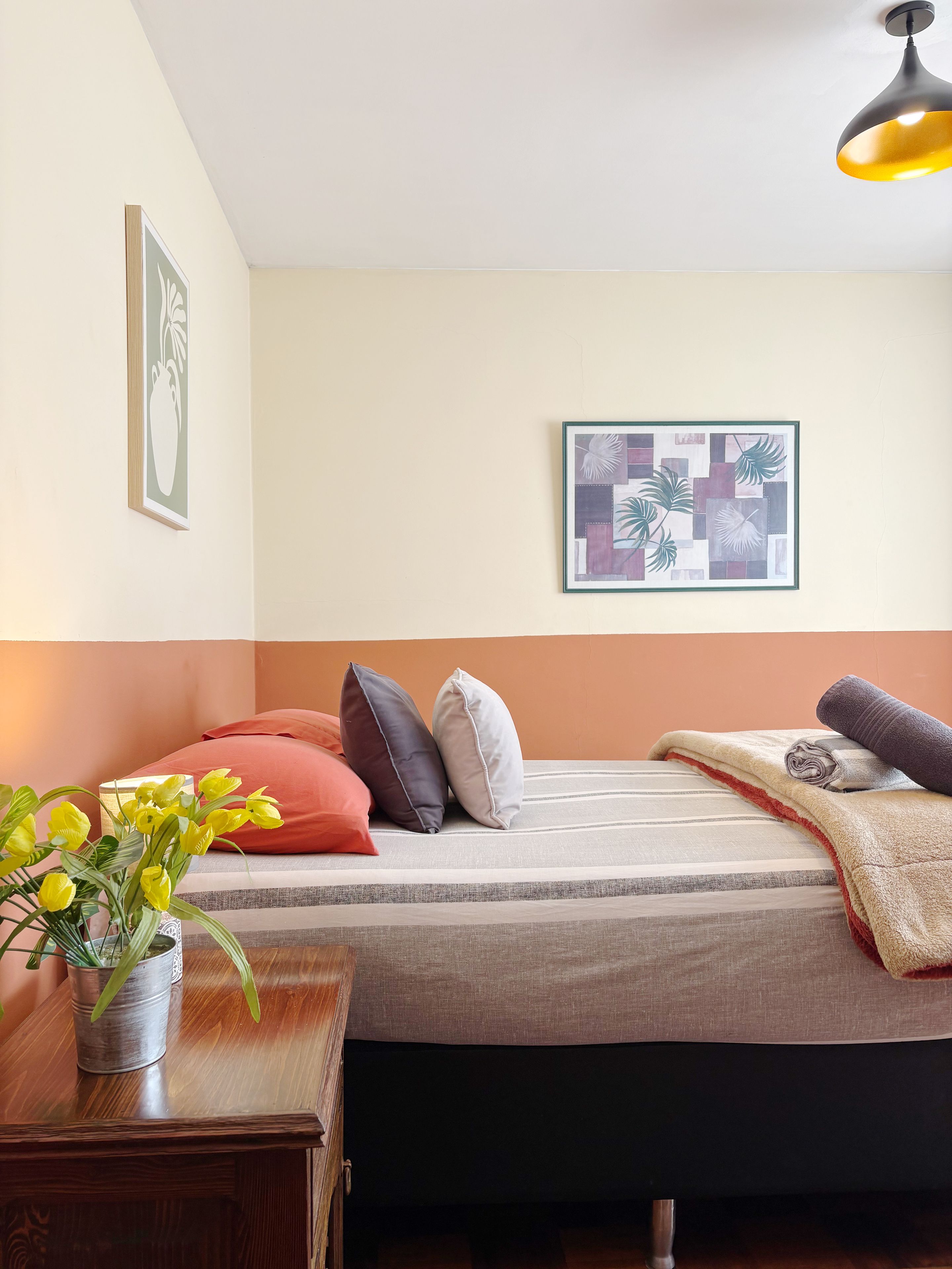 Quarto com cama, almofadas, manta, mesa de cabeceira com um vaso e obras de arte na parede.