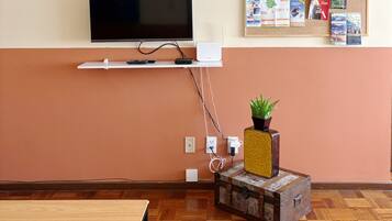 A room with a flat-screen TV mounted on a wall, a bulletin board with various papers and a small potted plant on a decorative box, and a wooden table with a rug.