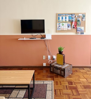 Quarto com televisão de tela plana na parede, um mural com diversos papéis e uma pequena planta em um vaso decorativo, e uma mesa de madeira com tapete.