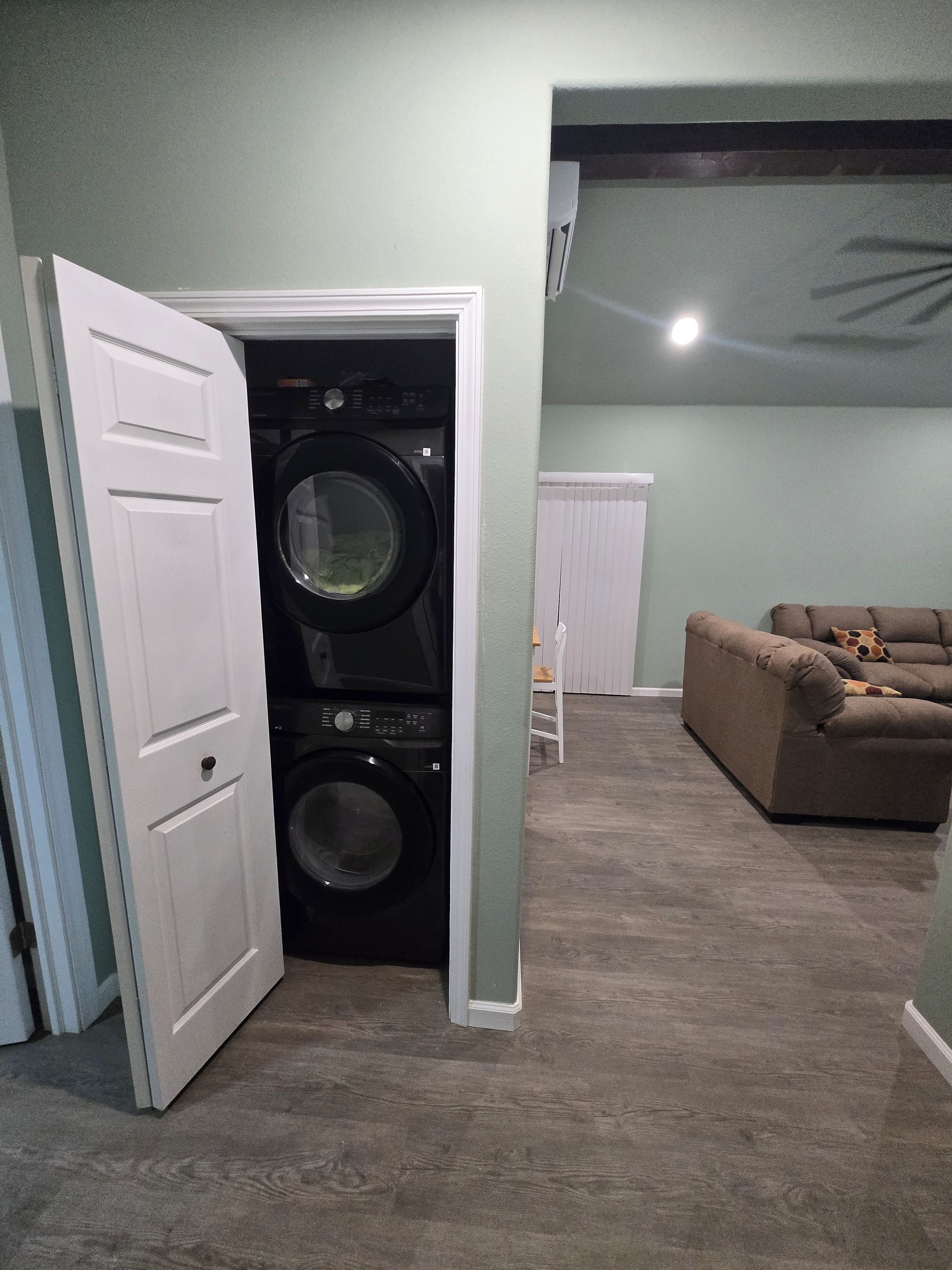 A laundry room with a front-loading washer and dryer, a white door, and a brown couch in the adjacent living area.