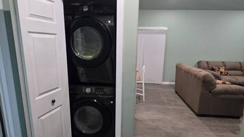 A laundry room with a front-loading washer and dryer, a white door, and a brown couch in the adjacent living area.