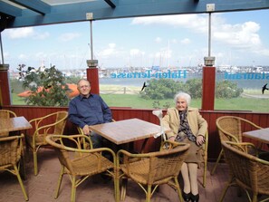 Two people seated at a table with wicker chairs, overlooking a marina with boats and cranes. - "studio" in the "white House Varwig" (Nebel)
