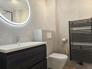 Ein modernes Badezimmer mit einem runden Spiegel, einem weißen Waschbecken, einer Toilette und einem Handtuchhalter.