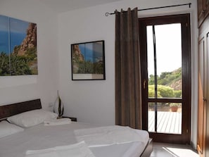 A bedroom with a bed, a nightstand, a picture frame, a window with curtains, and a view of greenery outside. - 1-bedroom Villa (Trinità d'Agultu e Vignola)