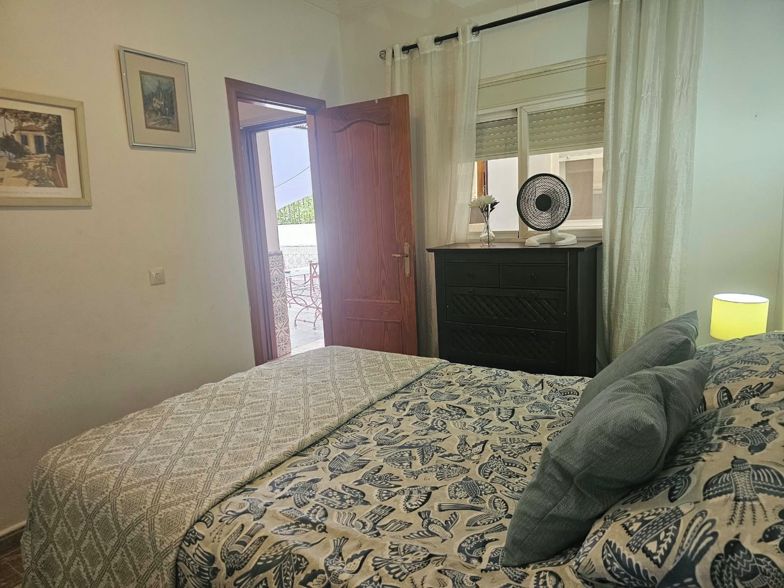 Un dormitorio con cama, cómoda, ventilador y ventana con cortinas.