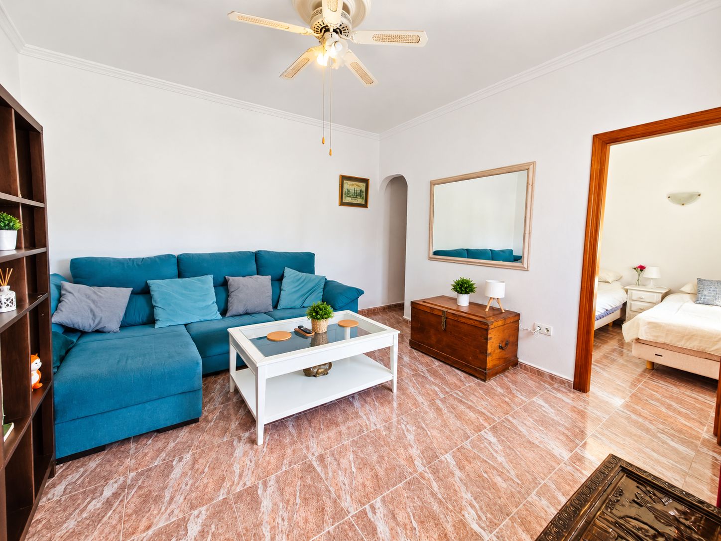 Una sala de estar con un sofá azul, una mesa de centro blanca, un aparador de madera y un ventilador de techo.