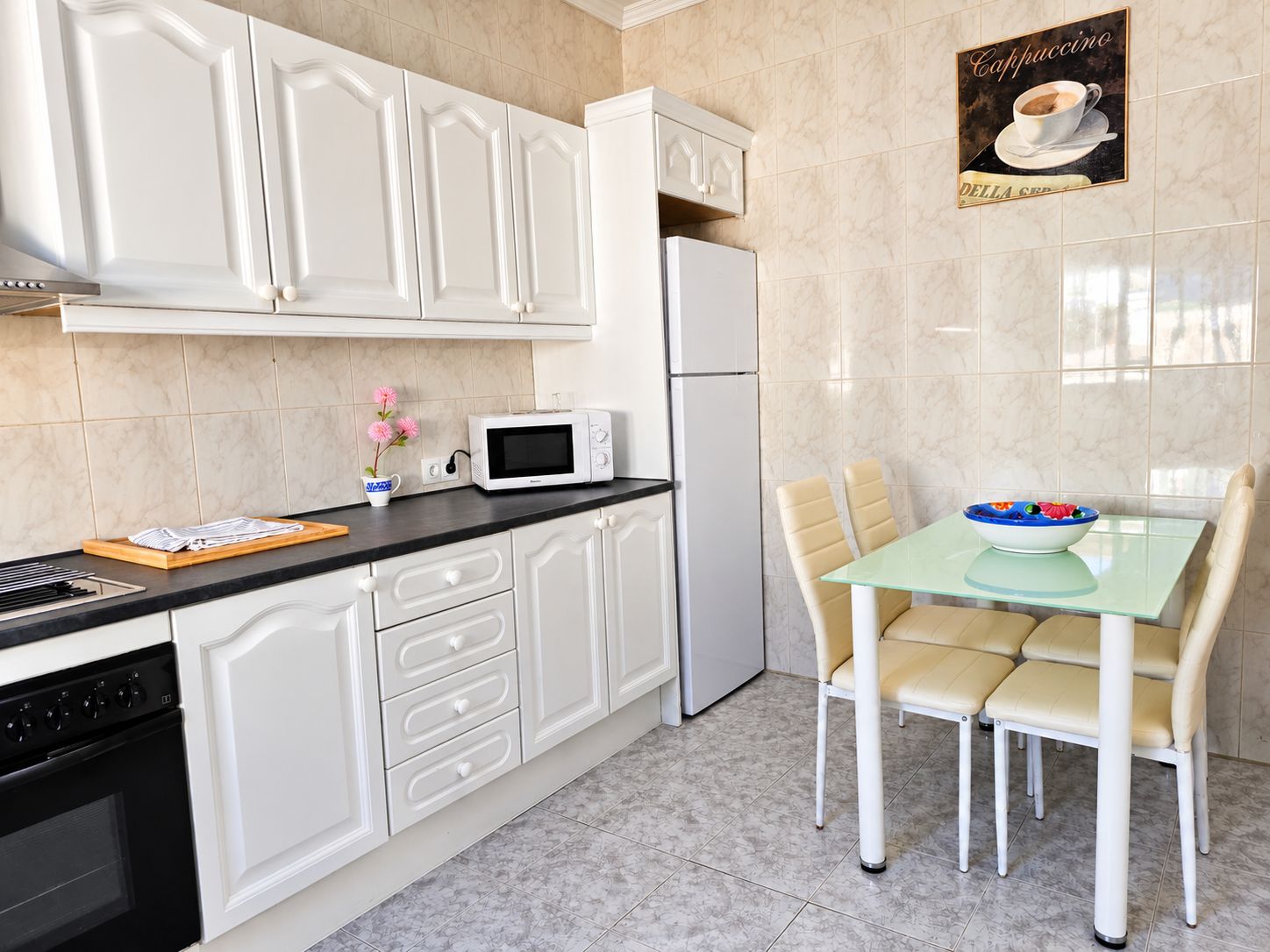 Een moderne keuken met witte kasten, een magnetron, een koelkast, en een eethoek met een glazen tafel en stoelen.