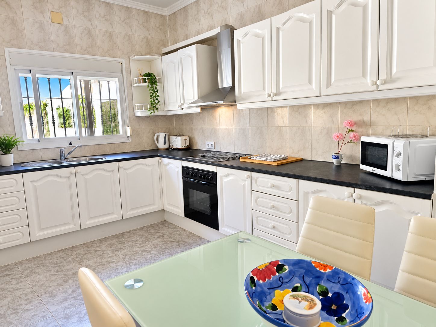 Una cocina moderna con armarios blancos, estufa negra, microondas y una mesa de comedor de vidrio con un cuenco floral.