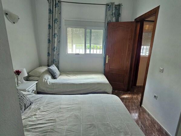 A bedroom with two beds, a window with blinds, a nightstand with a lamp, and a wooden door.