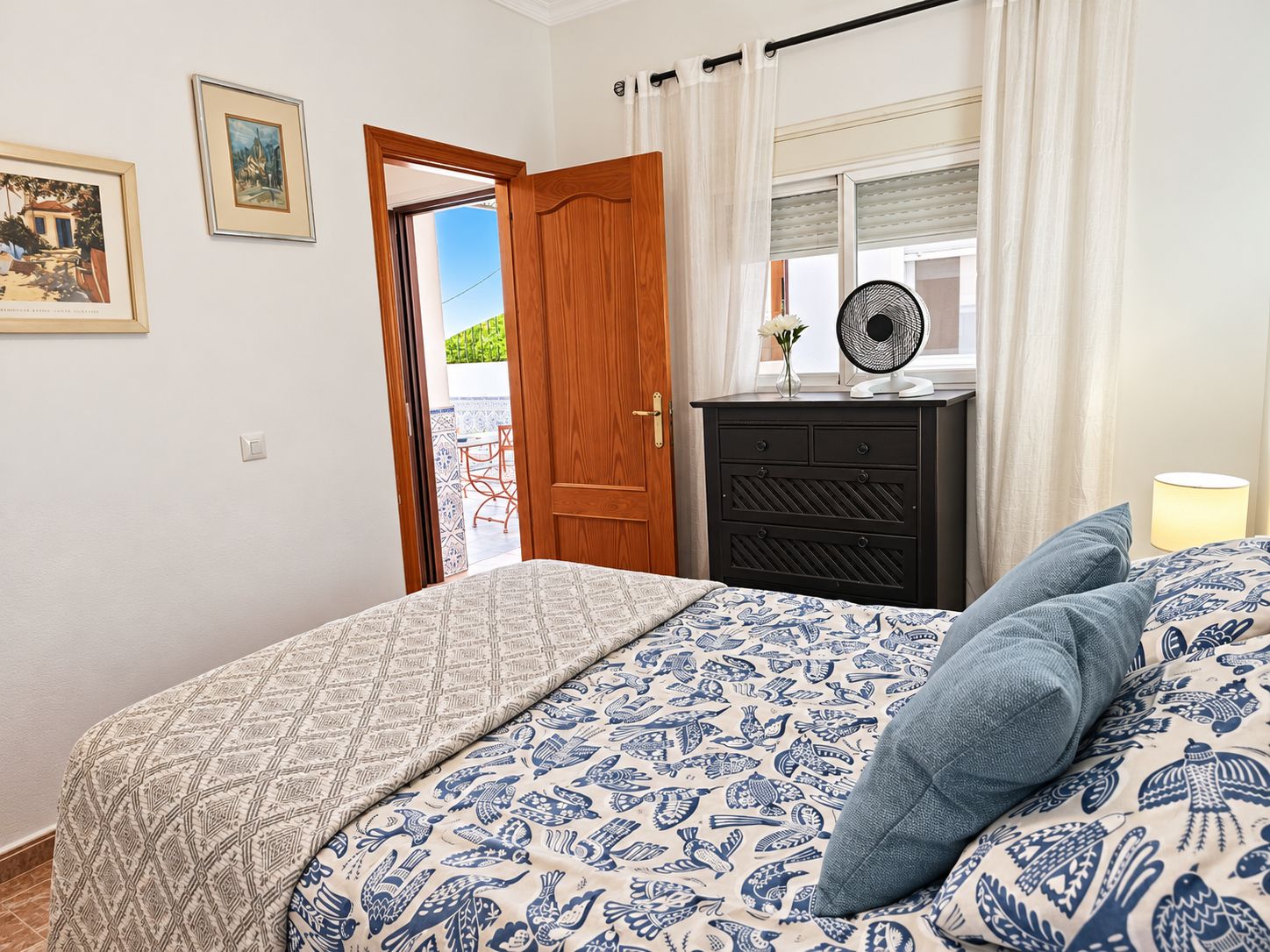 Een slaapkamer met een bed, een dressoir, een ventilator en een raam met gordijnen.