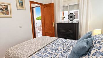 Een slaapkamer met een bed, een dressoir, een ventilator en een raam met gordijnen.