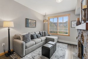 Ein modernes Wohnzimmer mit einem Ecksofa, einer Stehlampe, einem großen Fenster mit Jalousien und einer rustikalen Steinwand.