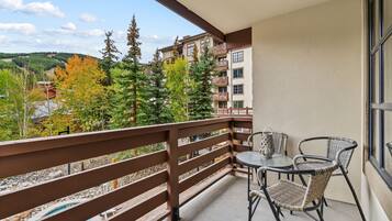 A balcony with a wooden railing, a round glass table, two wicker chairs, and a view of a forested area with autumn-colored trees.