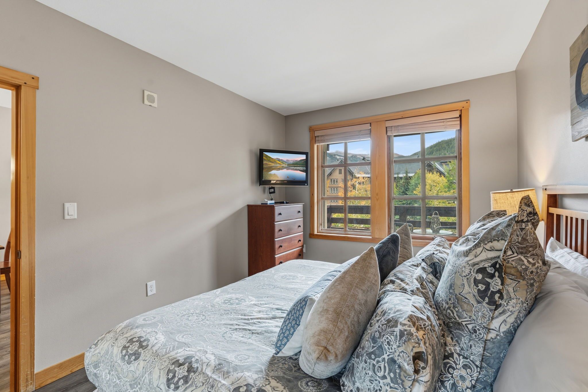 Un dormitorio con cama, televisor, cómoda y ventana con vistas a una casa y árboles.