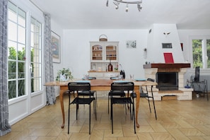 A dining area with a wooden table and chairs, a fireplace, and a window with patterned curtains. - Loargann ker Pondi - Maison pour 6 (Pontivy)