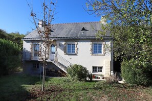 Une maison en pierre à deux étages, dotée d’un balcon, d’une cheminée et d’un petit jardin.