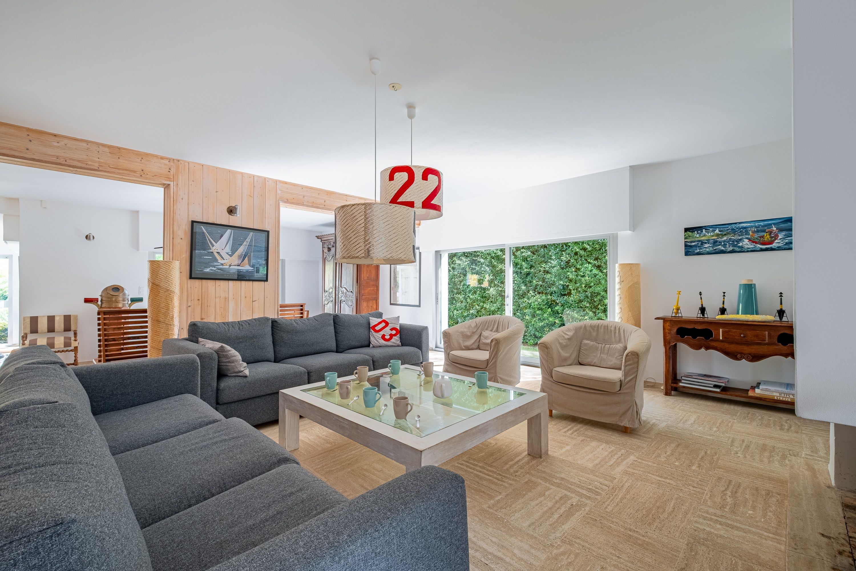 Un salon moderne avec un canapé gris, une table basse et un plancher en bois. Un tableau orne le mur et une fenêtre offre une vue sur la verdure.