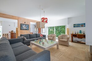 A modern living room with a grey sofa set, a coffee table, and a wooden floor. There is a painting on the wall and a window with a view of greenery. - Ker-eole - A côté du Port de la Trinité (La Trinite-sur-Mer)
