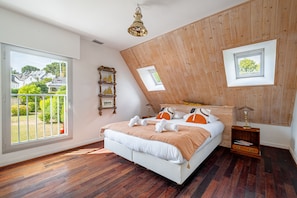 A bedroom with a sloped ceiling, a bed with white bedding, a wooden headboard, a bedside table with a lamp, and a window with a view of greenery. - Ker-eole - A côté du Port de la Trinité (La Trinite-sur-Mer)