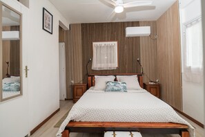Ein Schlafzimmer mit einem Holzkopfteil, einem Bett mit gemusterten Betttextilien, einem Nachttisch, einem Schreibtisch und einem Deckenventilator, sowie einem Fenster mit durchscheinendem Vorhang.