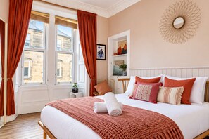 Ein Hotelzimmer mit einem großen Bett, roten Vorhängen, einem weißen Kopfteil, einem wandmontierten Spiegel und Blick auf die Stadt durch das Fenster.