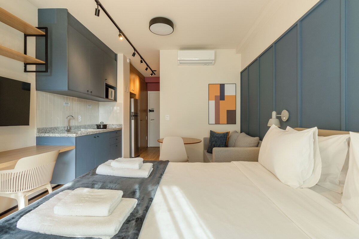 A modern hotel room with a bed, a seating area, a kitchenette, and a bathroom.