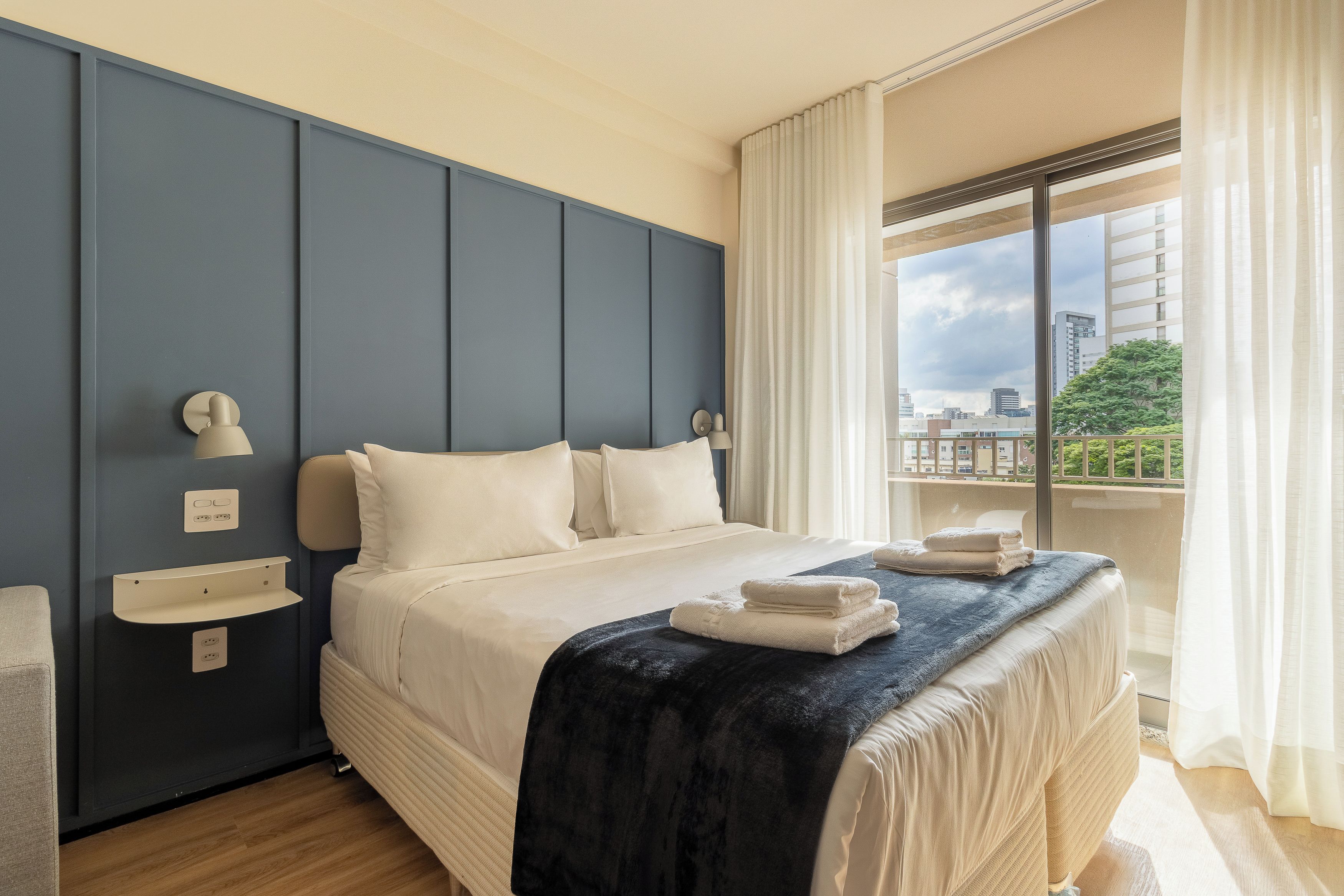 A hotel room with a large bed, a bedside table, a window with curtains, and a view of the cityscape.