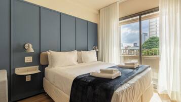 A hotel room with a large bed, a bedside table, a window with curtains, and a view of the cityscape.