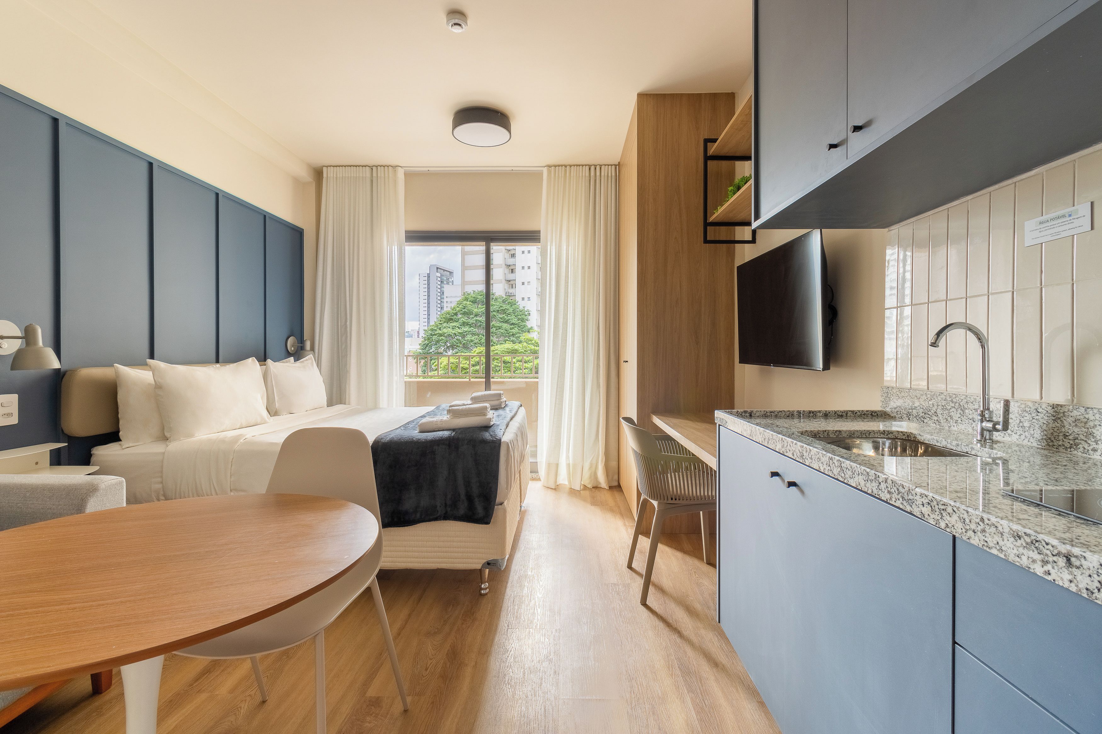 Una habitación de hotel moderna con cama, zona de comedor, cocina con lavabo y televisor montado en la pared.