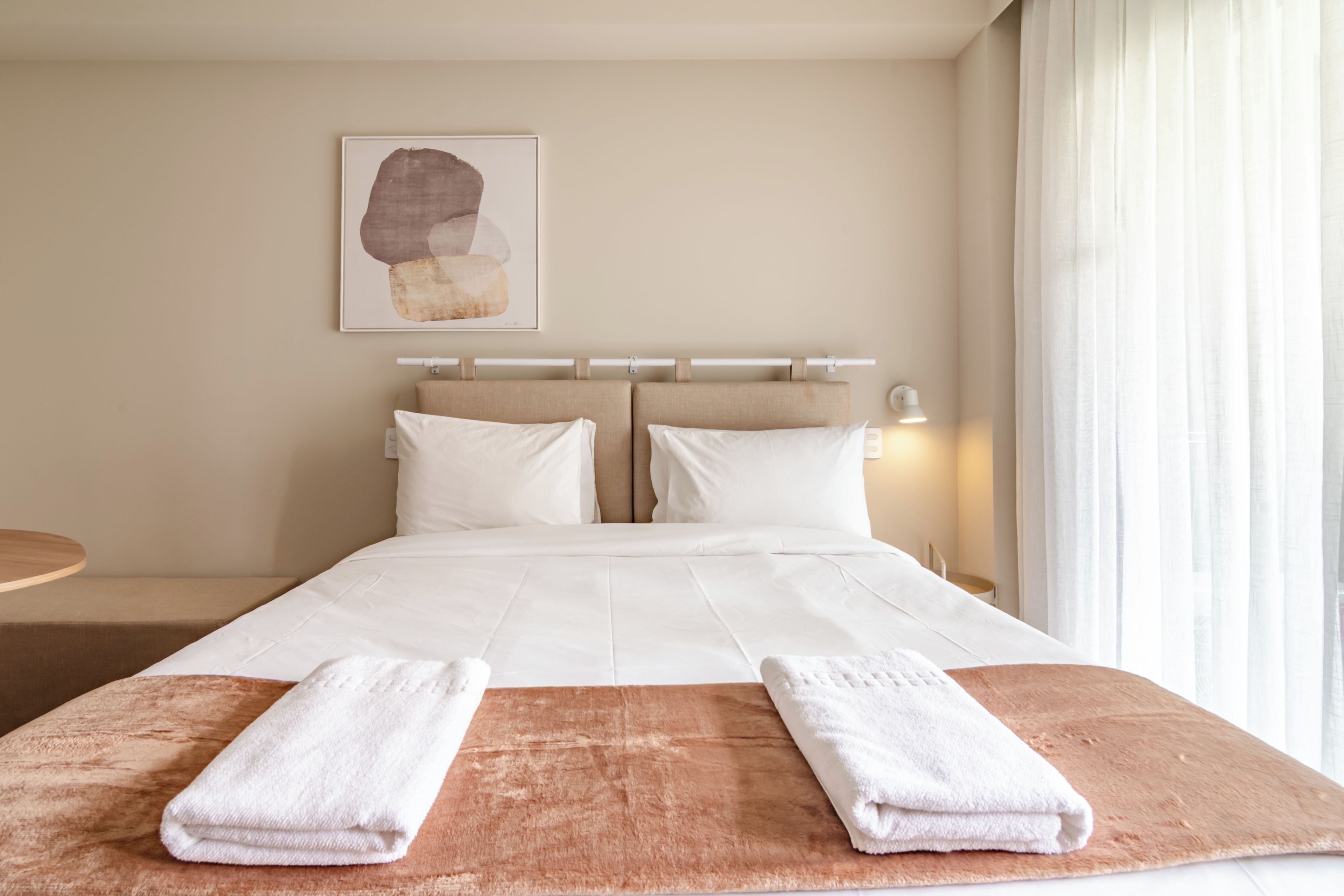 Habitación de hotel con cama, dos almohadas, cabecero, mesita de noche, lámpara y ventana con cortinas.