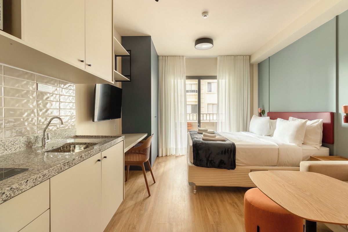 A hotel room with a bed, a desk, a chair, a TV, and a bathroom with a sink and mirror.