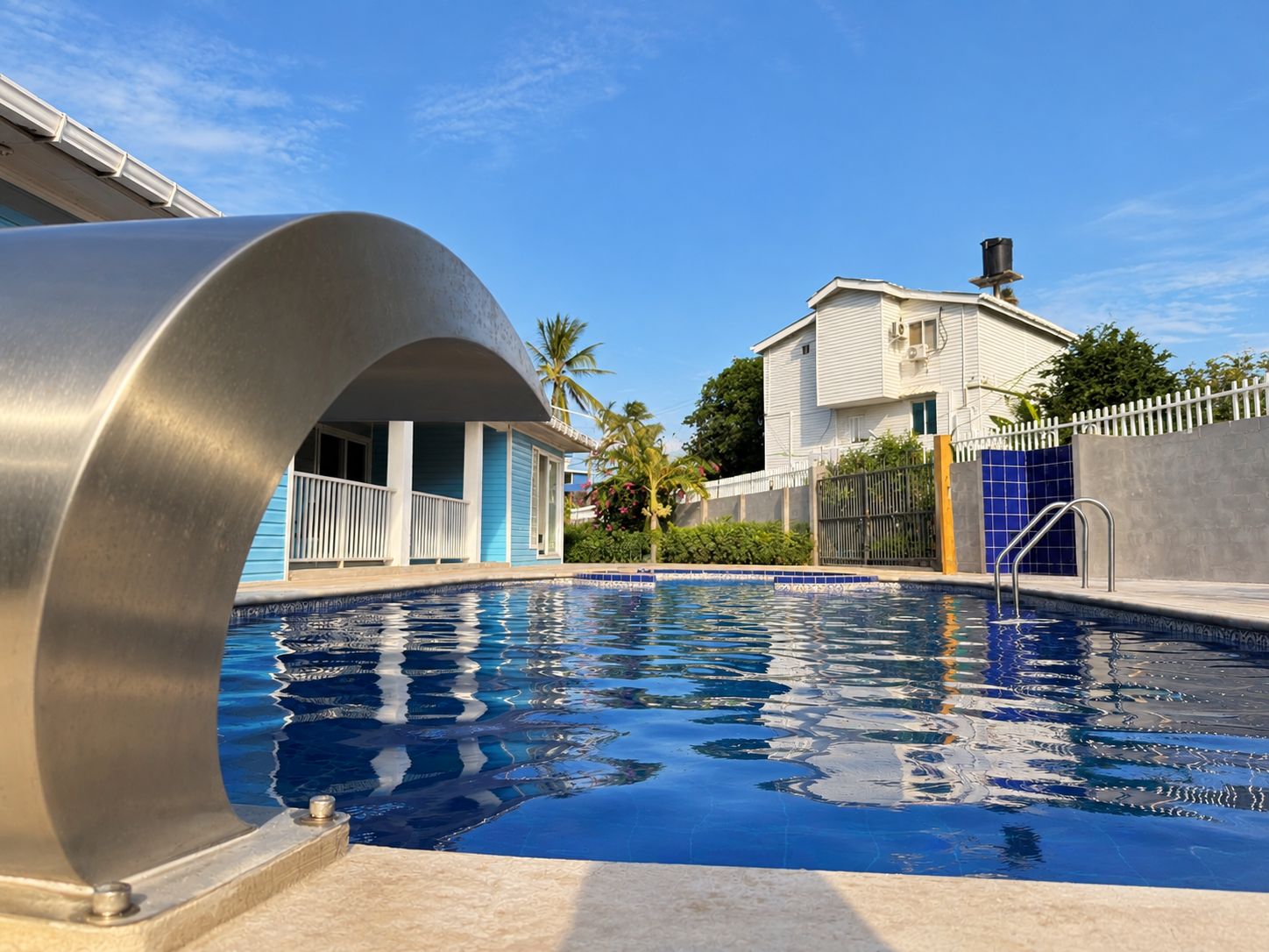 Une piscine à l’eau d’un bleu limpide, entourée d’une clôture blanche, d’une maison bleue et d’un palmier.