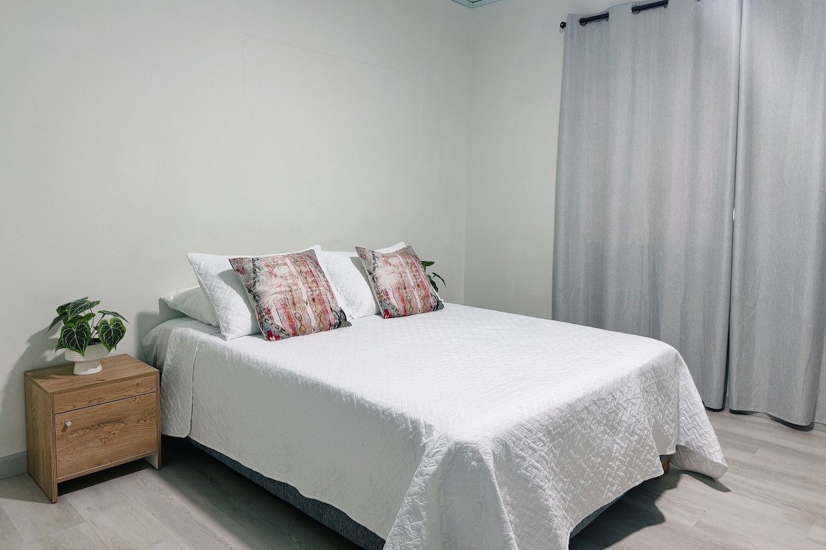 A bedroom with a bed, a nightstand, a potted plant, and a window with curtains.