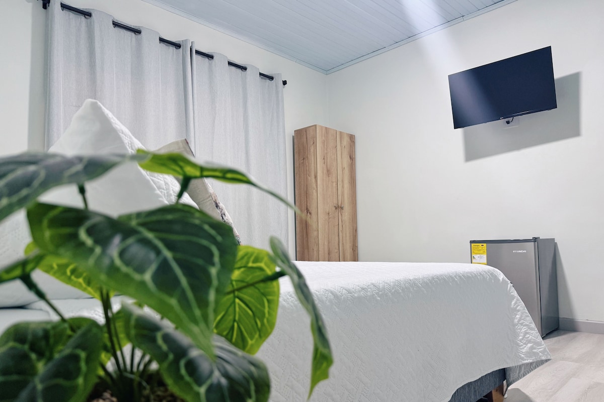 A hotel room with a bed, a television mounted on the wall, a wooden wardrobe, and a small refrigerator.
