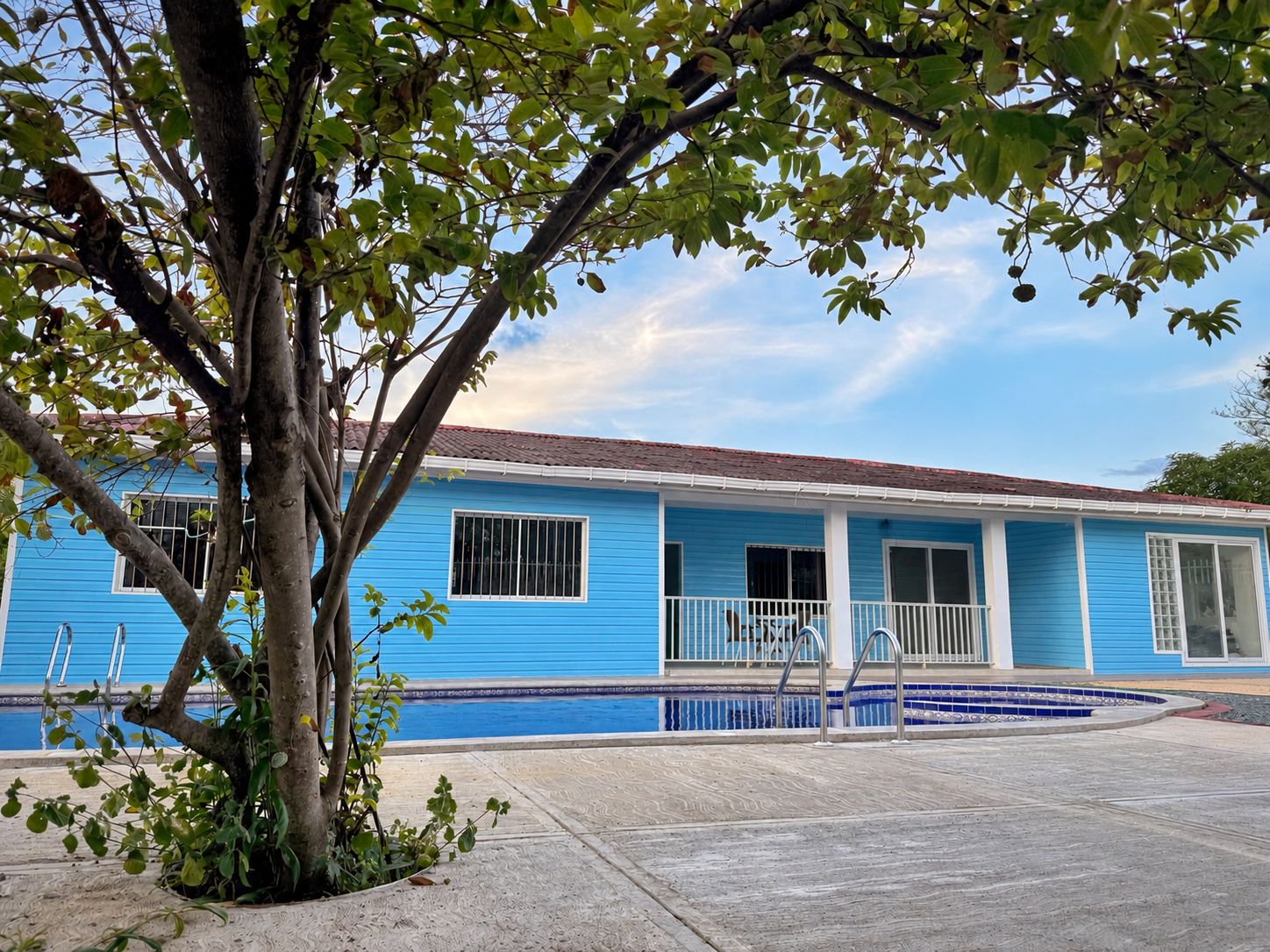 Une maison bleue avec une piscine, un arbre devant et un ciel dégagé.