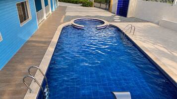 A rectangular swimming pool with a tiled edge, surrounded by a concrete deck, and adjacent to a blue building with a tiled roof.
