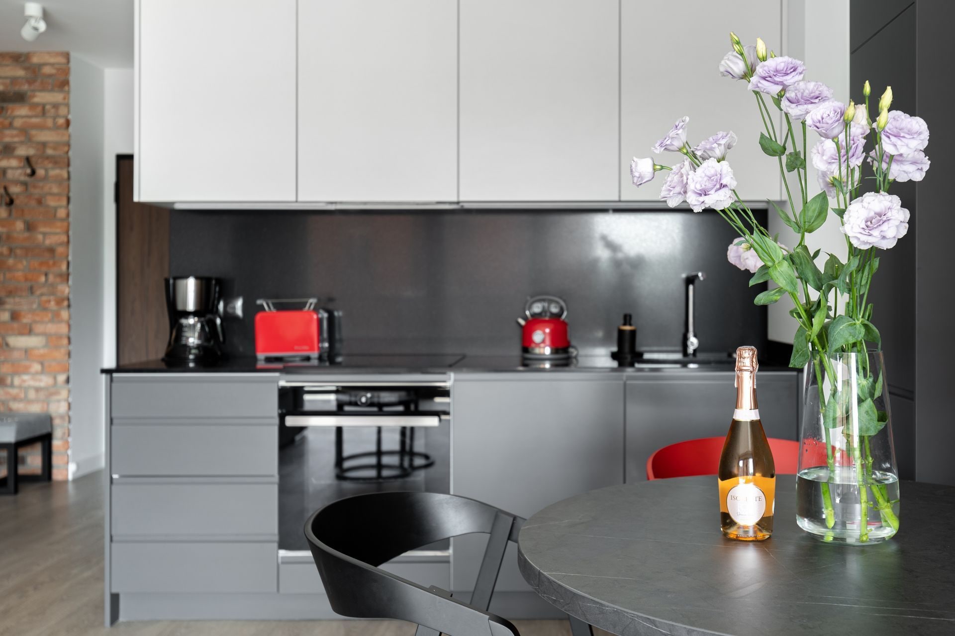 A modern kitchen with grey cabinets, a red kettle, and a table with a bottle and flowers.