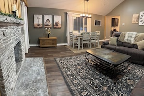 A living room with a stone fireplace, a dining area with a table and chairs, and a sofa with a plaid throw. - Long Term Fully Furnished Country Farmhouse Ranch w/ pond in Rock Creek, Ohio (Rock Creek)