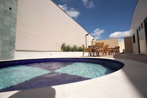 Ein Pool mit geschwungener Kante, umgeben von einer beigen Wand und einer gefliesten Fläche. In der Nähe steht ein Holztisch mit Stühlen.