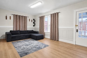 A modern living room with a sectional sofa, a patterned rug, and a door with a view of buildings. - 2BR Lower Unit Near Niagara Falls | Free WiFi (Niagara Falls)