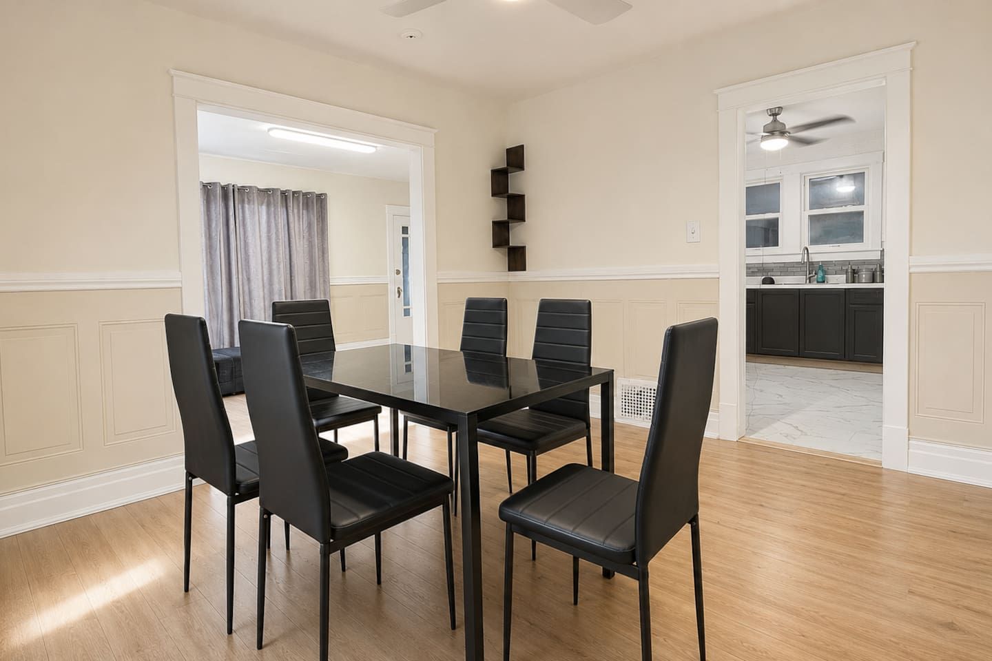 A dining area with a rectangular table and six chairs, a kitchen in the background, and a ceiling fan.