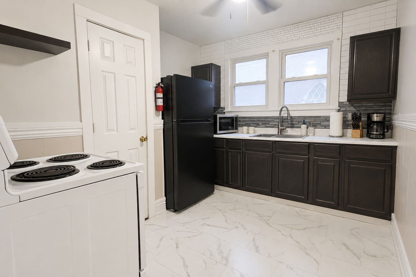 A kitchen with a white stove, black refrigerator, dark cabinets, a white countertop, and a window with blinds.