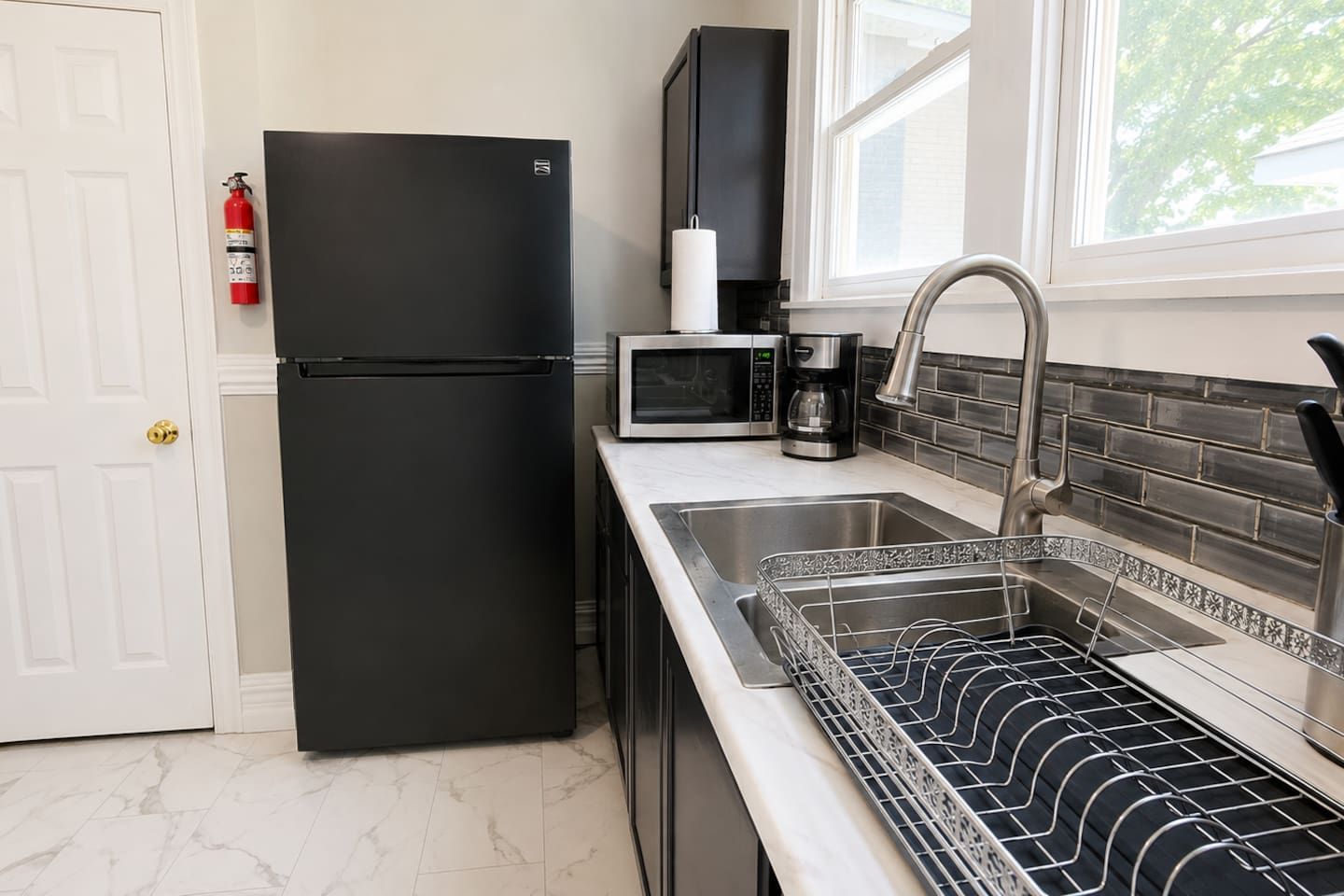 A kitchen with a black refrigerator, a microwave, a coffee maker, and a stainless steel sink with a dish rack.
